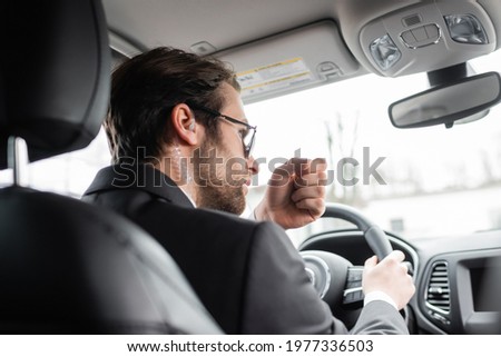 bearded bodyguard in sunglasses and security earpiece talking while driving modern automobile