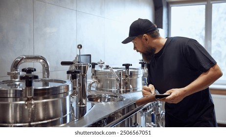 Bearded brewery technologist in black cap carefully inspects stainless steel brewing equipment, taking notes on clipboard. Craftsmanship of beer production. Concept of beer, brewery, quality control - Powered by Shutterstock