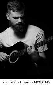 Bearded Brutal Guitarist Plays An Acoustic Guitar In A Black Room. The Concept Of Music Recording, Rehearsal Or Live Performance.