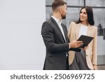 Bearded business man car salesman in grey suit giving explanations on tablet to pretty young woman in beige jacket outside car dealership