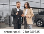 Bearded business man car salesman in grey suit giving explanations on tablet to pretty young woman in beige jacket outside car dealership