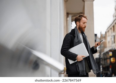 Bearded business man in a jacket and scarf with a laptop using a smartphone while standing on the street near the entrance in the building - Shutterstock ID 2267847569