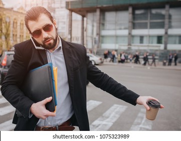Bearded businessman with coffee cup catching a taxi. He does not have time, he is going to talk on the phone on the go. Man doing multiple tasks. Multitasking business male persone. - Shutterstock ID 1075323989