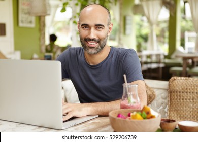 Bearded businessman dressed casually checking email on his laptop during breakfast, sitting at nice cafe, drinking smoothie, looking at camera with happy confident face expression, enjoying vacations - Powered by Shutterstock