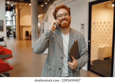 Bearded businessman in eyeglasses with laptop is talking phone standing in modern coworking space - Shutterstock ID 2395459407
