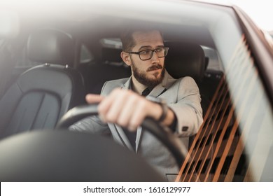 A bearded businessman with glasses sits behind the wheel of an Executive car and looks away. Photo through the windshield