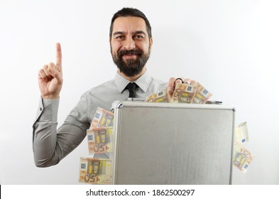 Bearded businessman on white background smiling with raised arm and pointing finger up holding up with his hand a briefcase full of Euro banknotes sticking out. Business concept