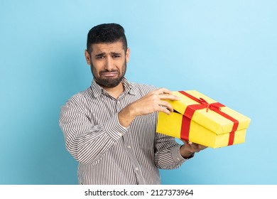 Bearded Businessman Opening Gift Box, Looking At Camera With Disappointed Sad Expression, Unwrapping Bad Present, Wearing Striped Shirt. Indoor Studio Shot Isolated On Blue Background.