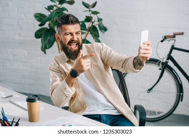 Bearded Businessman Pointing At Camera While Taking Selfie In Office Chair