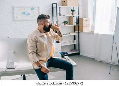 bearded businessman sitting on office desk and drinking coffee from disposable cup 