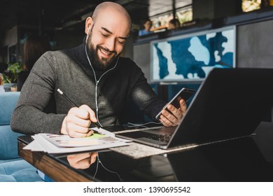 Bearded businessman is working on laptop, making notes in document, speaks in headphones on cell phone while sitting at table in coffee shop.Young hipster uses smartphone and computer.Online education