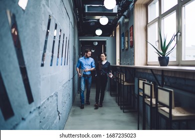 Bearded Casual Man And Formal Woman Walking Down Narrow Hallway With Counter Alongside Window And Chatting About Work Issues In Modern Office Building