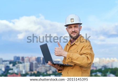 Bearded caucasian chief engineer or construction architect in white helmet and orange jacket holding laptop in front of urban city background. Making thumbs up gesture, smiling and looking at camera