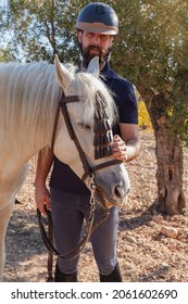 A Bearded Caucasian Man In Equestrian Clothing And Helmet Is Standing Still Stroking The Head Of His White Horse In A Field With Olive Trees.