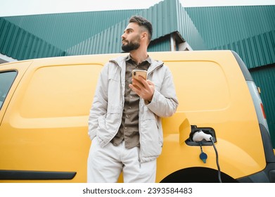 bearded caucasian man standing near an electric car that is charging and making time adjustments on a smartphone - Powered by Shutterstock