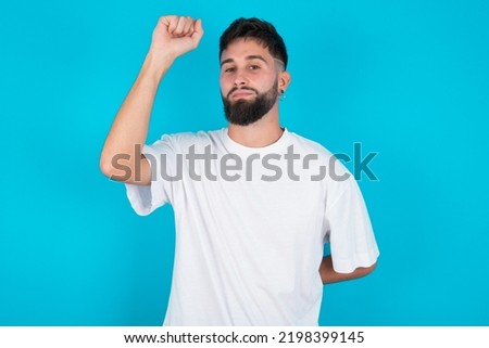 bearded caucasian man wearing white T-shirt over blue background feeling serious, strong and rebellious, raising fist up, protesting or fighting for revolution.
