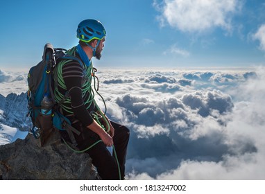 Bearded Climber In A Safety Harness, Helmet, And On Body Wrapped Climbing Rope With Sitting At 3600m Altitude On A Cliff And Looking At  Picturesque Clouds During Mont Blanc Ascending, France Route