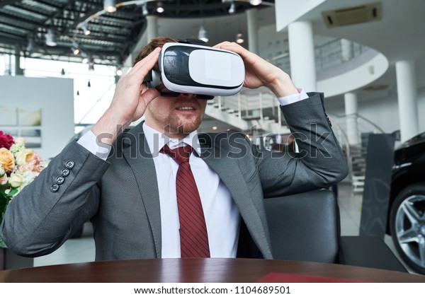 Bearded customer wearing\
classical suit sitting at wooden table and using VR headset while\
undergoing virtual test-drive, interior of modern car showroom on\
background