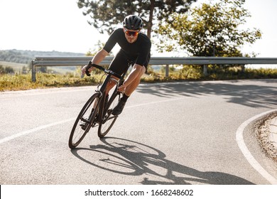 Bearded cyclist in sport clothing, protective helmet and mirrored glasses riding black bike among countryside nature during summer time. Mature man preparing for competitions and races.