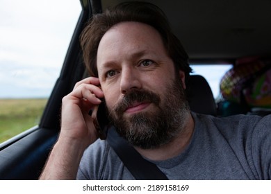 A Bearded Dad Looks Out The Window Of A Car While Talking On A Smart Phone On A Road Trip On Summer Vacation.  Lifestyle Image Of A Candid Moment Of This Middle Aged Man.