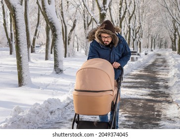 A Bearded Dad Walking With Baby Carriage In Winter Park. A Man With A Baby Stroller Walks In A Winter Snow-covered City Park In The Cold. Caring Father Admires Baby Sleeping In Stroller