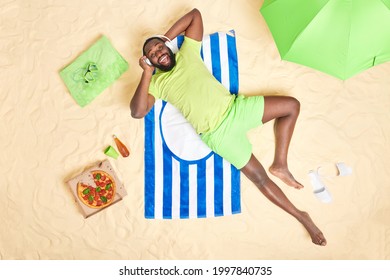 Bearded dark skinned Afro American man wears casual green t shirt shorts listens music via headphones lies on blue striped towel poses near pizza slippers parasol has fun. Vacation at seaside. - Powered by Shutterstock