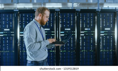 Bearded Data Center IT Engineer in Glasses is Walking Through Server Rack Corridor with a Laptop Computer. He is Visually Inspecting Working Server Cabinets.