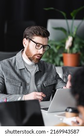 Bearded Designer In Eyeglasses Working Near Laptop In Ad Agency Near Blurred Colleague