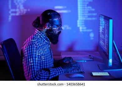 Bearded developer in eyeglasses sitting at the table in front of computer monitor typing on computer keyboard and developing new software