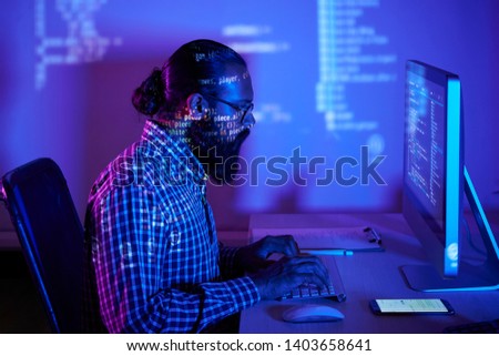 Bearded developer in eyeglasses sitting at the table in front of computer monitor typing on computer keyboard and developing new software