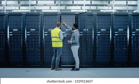 Bearded IT Engineer in Glasses and High Visibility Vest with a Laptop Computer and Black Technician Colleague Talking in Data Center while Standing Next to Server Racks. Running Maintenance. - Powered by Shutterstock