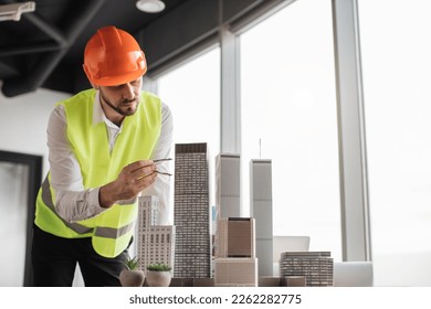 Bearded engineer in protective helmet working on building complex prototype project of residential or business district, measuring city model using compass for drawing at office with panoramic view - Powered by Shutterstock