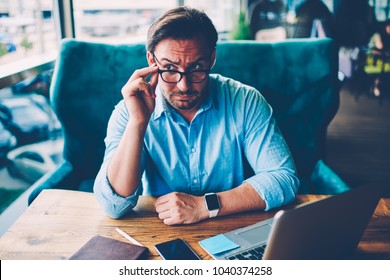 Bearded entrepreneur dressed in blue shirt fixing optical eyeglasses with black frame while looking away sitting at wooden desktop with digital laptop computer connected to wireless internet - Shutterstock ID 1040374258