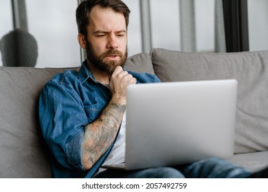 Bearded European Man Working With Laptop While Sitting On Sofa At Home