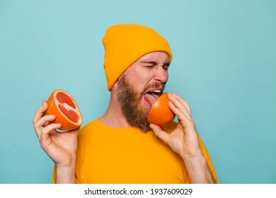 Bearded european man in yellow shirt isolated on turquoise background holding grapefruit tastes, wrinkles from bitterness