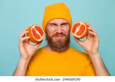 Bearded european man in yellow shirt isolated on turquoise background holding grapefruit tastes, wrinkles from bitterness