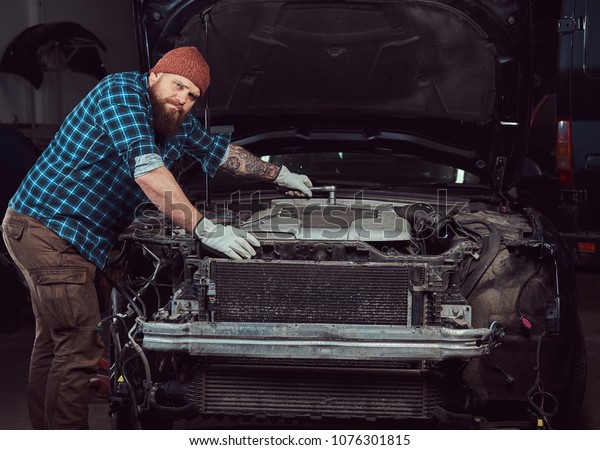 A bearded expert\
mechanic dressed in a uniform, standing with crossed arms against a\
car in the garage.