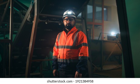 Bearded factory worker in uniform and hardhat looking at camera while standing under conveyor belt in dim workshop