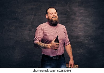 A bearded fat male dressed in a pink fleece shirt holds a beer bottle over dark grey background.