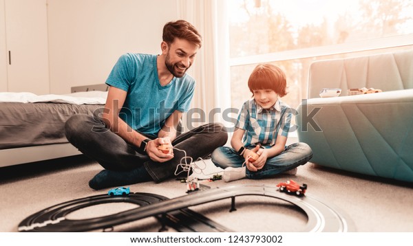 Bearded Father\
and Son Playing with Toy Race Road. Man Sitting on Floor. White\
Carpet in Room. Toy Cars. Exited Boy. Happy Family Concept. White\
Carpet. Lying on Floor. Indoor\
Fun.