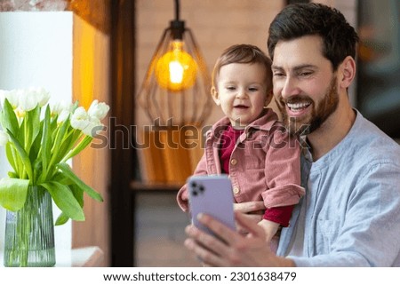 Bearded father standing with baby daughter and mobile phone having video call or selfie