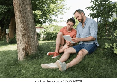 bearded father and teenager son looking at digital tablet while sitting near tree in park - Powered by Shutterstock