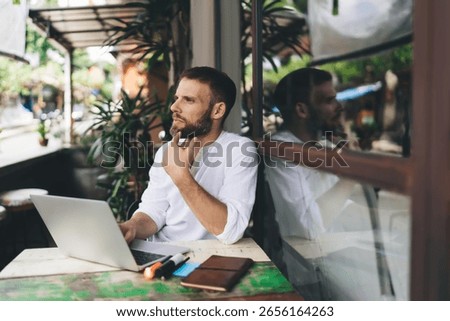 Bearded freelancer pausing in reflection, touching chin, looking away from screen. Scene illustrates conscious tech use, digital mindfulness and self-direction in modern location-flexible work life.