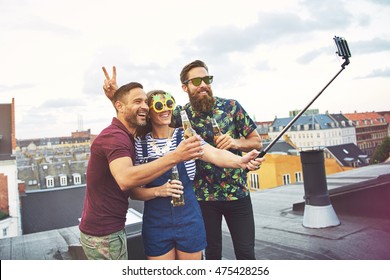 Bearded friend giving rabbit ears to man with beer while woman in sunglasses holds a camera phone at the end of a selfie stick - Shutterstock ID 475428256