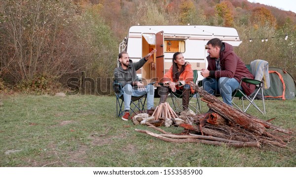 Bearded friend\
telling a story to his couple friends in front of camper van. Wood\
for fire. Friends\
relaxing.