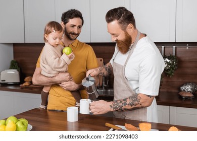 Bearded gay man pouring coffee near husband holding toddler daughter in kitchen - Powered by Shutterstock