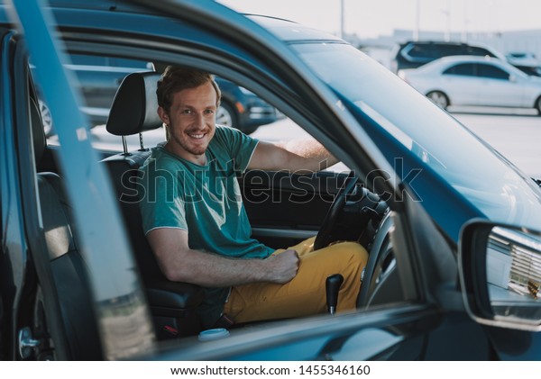 Bearded gentleman sitting in the\
driver seat inside automobile. He looking at camera and\
smiling