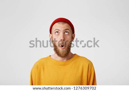 The bearded guy looks at the top with surprise, mouth rounded. Portrait of a man in a red cap looking at the empty copy space from above, wants to say Wow. That's so unexpected.