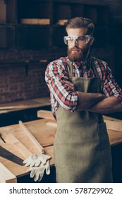 Bearded handsome joiner  think and look to the side near tabletop with tools.  Stylish young entrepreneur with brutal hairstyle and saved glasses crossed hands at his workstation.