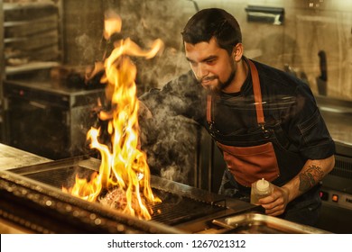 Bearded handsome man in jeans apron and with tattoo on hand cooking. Chef smiling, holding oil in hand and looking at meat on grill. Tasty beef steak flaming with fire and smoke.  - Powered by Shutterstock